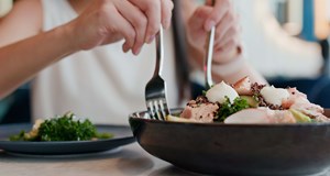 A person tucking into a healthy bowl of salad with a knife and fork. The rest of the table contains another plate with leafy greens.