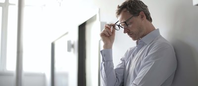 Person wearing a shirt leaning against a wall looking down to the ground with a worried expression 