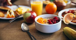 A white bowl full of fruit with a spoon next to it sitting on a table. The rest of the table contains more fruit and other healthy breakfast foods  