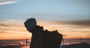 A homeless person with their hood up and rucksack on walking at dusk