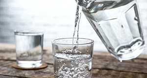 A small glass being filled with water from a clear water jug. The glass is sitting on a brown coaster on a wooden table, with another in the background.