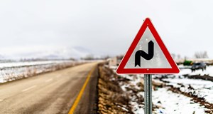 A road warning sign showing bends ahead next to a road with snow covered fields on either side
