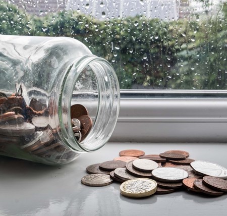 A jar full of money on its side with some of the coins spilled out on a windowsill on a rainy day
