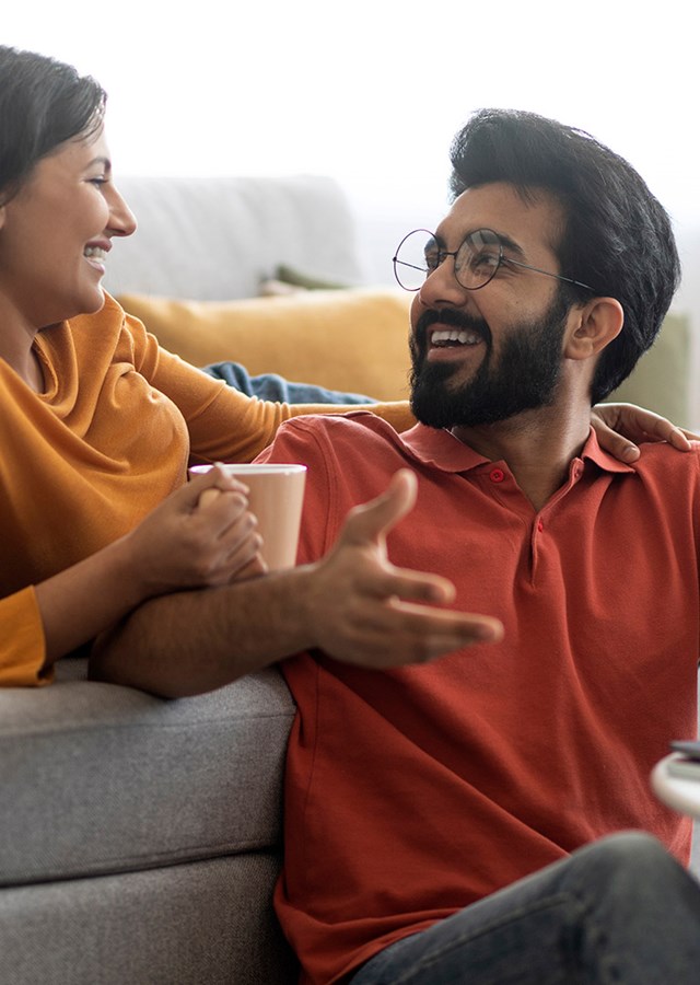 Happy couple discussing finances together on grey sofa with a laptop and coffee at home.