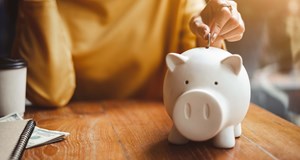 Person saving money and putting coins into a piggy bank with a notebook on the side working out their living costs