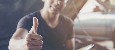 A person perched on an open car bonnet in a workshop smiling doing a thumbs up sign