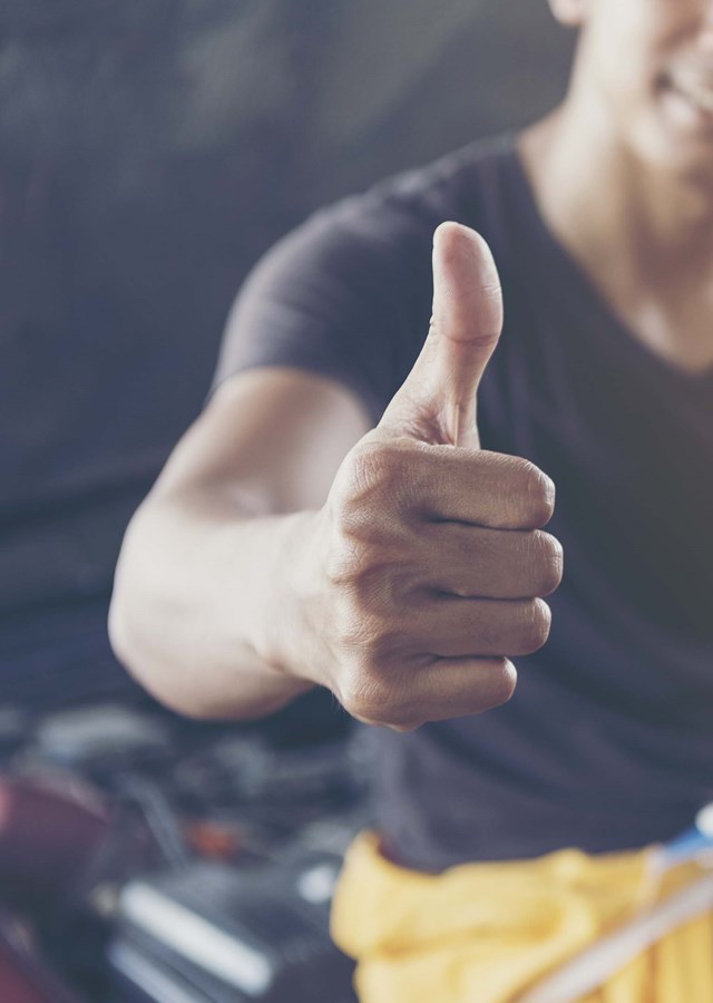A person perched on an open car bonnet in a workshop smiling doing a thumbs up sign