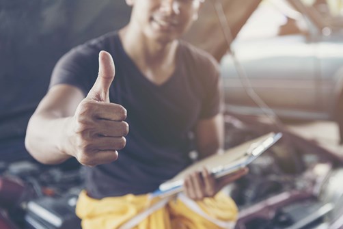 A person perched on an open car bonnet in a workshop smiling doing a thumbs up sign